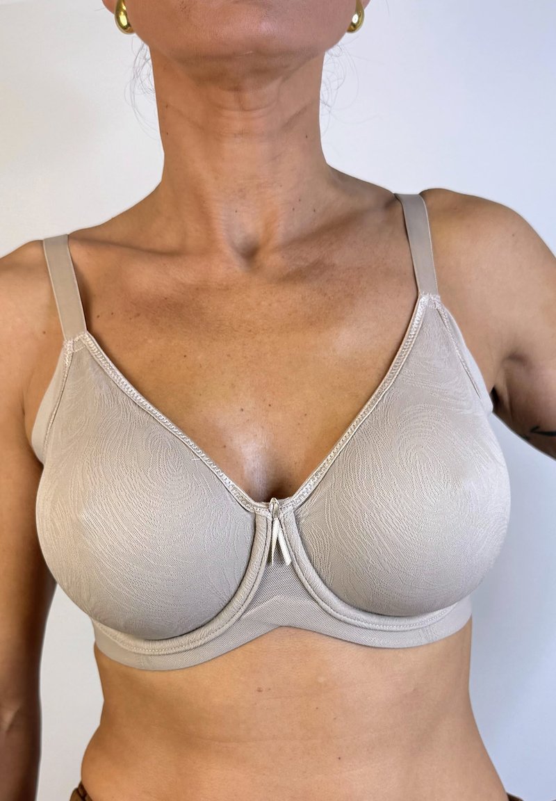 Sostén de color beige con un estampado, tirantes ajustables, copas suaves y un cierre frontal decorativo. Textura suave y bordes curvados son prominentes.