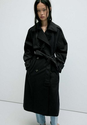 Frau mit langen schwarzen Haaren, die einen schwarzen Trenchcoat mit Gürtel und hellblaue Jeans trägt und vor einem schlichten hellgrauen Hintergrund steht.