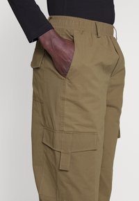 Khaki cargo kalhoty s elastickým pasem, bočními a náprsními kapsami. Tkanina má hladkou texturu a odolný vzhled.
