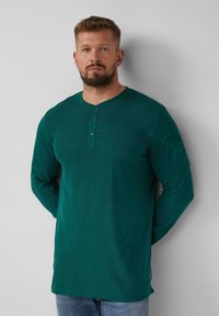 Langärmliges Henley-Shirt in dunkelblaugrün, mit Rundhalsausschnitt, einer Knopfleiste mit drei Knöpfen und einer Taschen auf dem Ärmel. Weicher, strukturierten Stoff.