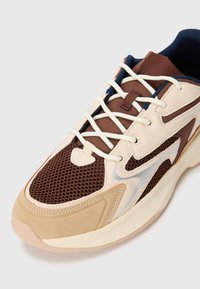 Beige og brun joggesko med meshstoff, semsket skinn-overflater, hvite snører og tykk polstret såle, fotografert på en hvit bakgrunn.