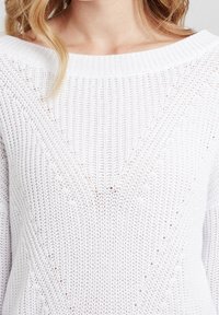 Biały sweter dzianinowy o otwartym splotcie, szerokim dekolcie w łódkę oraz teksturalnym trójkątnym wzorze z przodu, podkreślającym szczegóły ściegów.