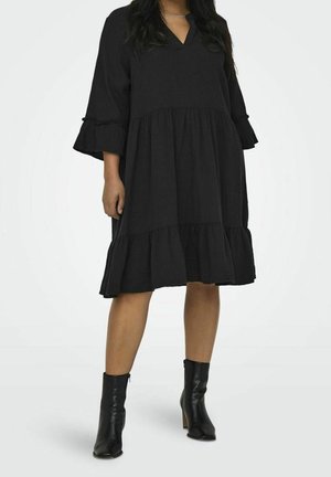 Frau trägt ein lockeres, schwarzes, knielanges Kleid mit Rüschen und schwarze Stiefeletten, steht vor einem weißen, einfarbigen Hintergrund.