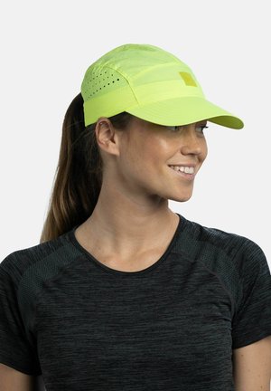 Junge Frau mit heller Haut, braunen Haaren im Pferdeschwanz, trägt eine leuchtend gelbe Sportmütze und ein schwarzes strukturiertes Sportshirt, lächelt und schaut nach rechts.