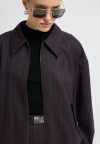 Donkerpaarse zip-up jas met streepdessin, brede kraag en zijzakken, gecombineerd met een zwart ribbeltrui en zilveren zonnebril.