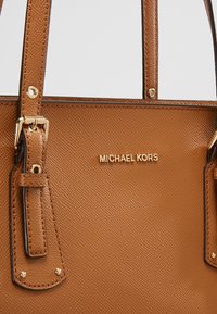 Sac à main Michael Kors en cuir marron texturé avec ferrures dorées et détail de bandoulière réglable sur un fond neutre.
