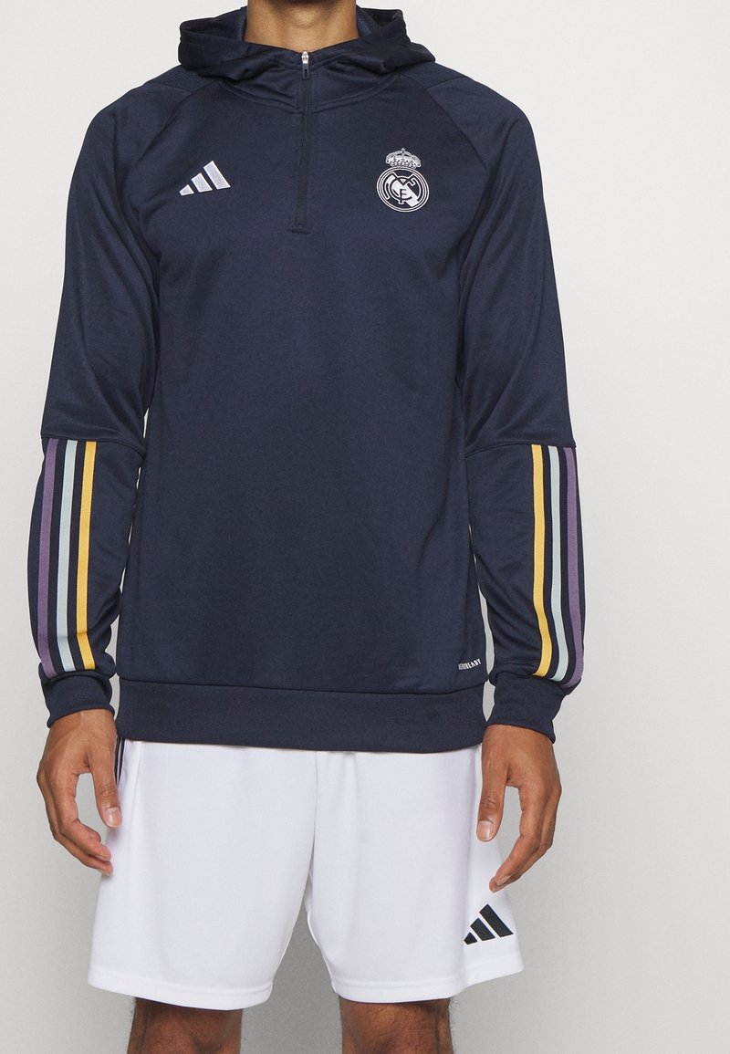 Sweat à capuche bleu marine avec demi-fermeture éclair, comportant le logo du Real Madrid et des rayures multicolores sur les manches. Fabriqué en tissu lisse.