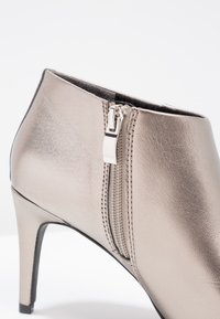 Botín de tobillo metálico en taupe con un diseño elegante, tacón alto de aguja, cremallera lateral y detalles de costura en material de cuero suave.