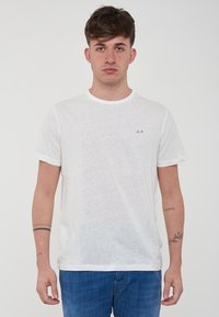 T-shirt bianco a maniche corte con collo rotondo, realizzato in tessuto leggero, con un piccolo logo blu sulla parte sinistra del petto.