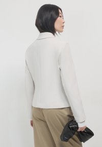 Una donna con capelli neri corti indossa un blazer bianco e pantaloni beige, tenendo una pochette in pelle nera su uno sfondo bianco semplice.