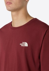 Camiseta de color burdeos hecha de algodón, con un diseño de cuello redondo y un pequeño logo blanco en la parte izquierda del pecho. Textura suave.