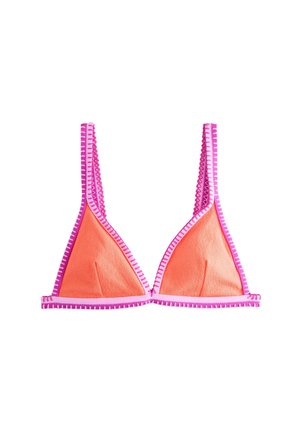 Mujer con el pelo largo y rizado, que lleva un bikini naranja con ribete rosa, de pie en una playa soleada con olas al fondo.