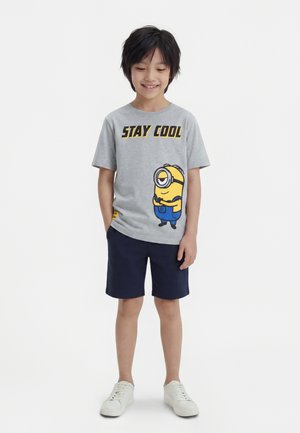 Garçon souriant portant un t-shirt gris avec le texte "Stay Cool" et un motif Minion, un short bleu marine et des baskets blanches, debout les mains dans les poches.