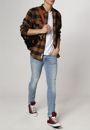 Chemise en flanelle à carreaux marron et noir portée sur un t-shirt blanc, jean skinny bleu clair, et baskets montantes bordeaux. Sac à épaule noir avec des accents.