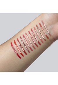 Campioni di colori per labbra mostrati su un braccio dalla pelle chiara. Dodici tonalità distinte, che variano dal nude al rosso, etichettate con nomi in testo bianco.