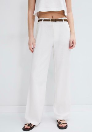 Žena nosi široke bijele hlače visokog struka s smeđim remenom, skraćeni bijeli top i crne sandale sa zlatnim kopčama na jednobojnoj pozadini.