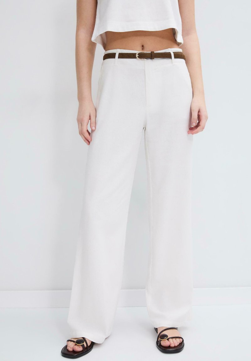 Femme portant un pantalon blanc taille haute à jambes larges avec une ceinture marron, un top blanc court et des sandales noires avec boucles dorées sur un fond uni.