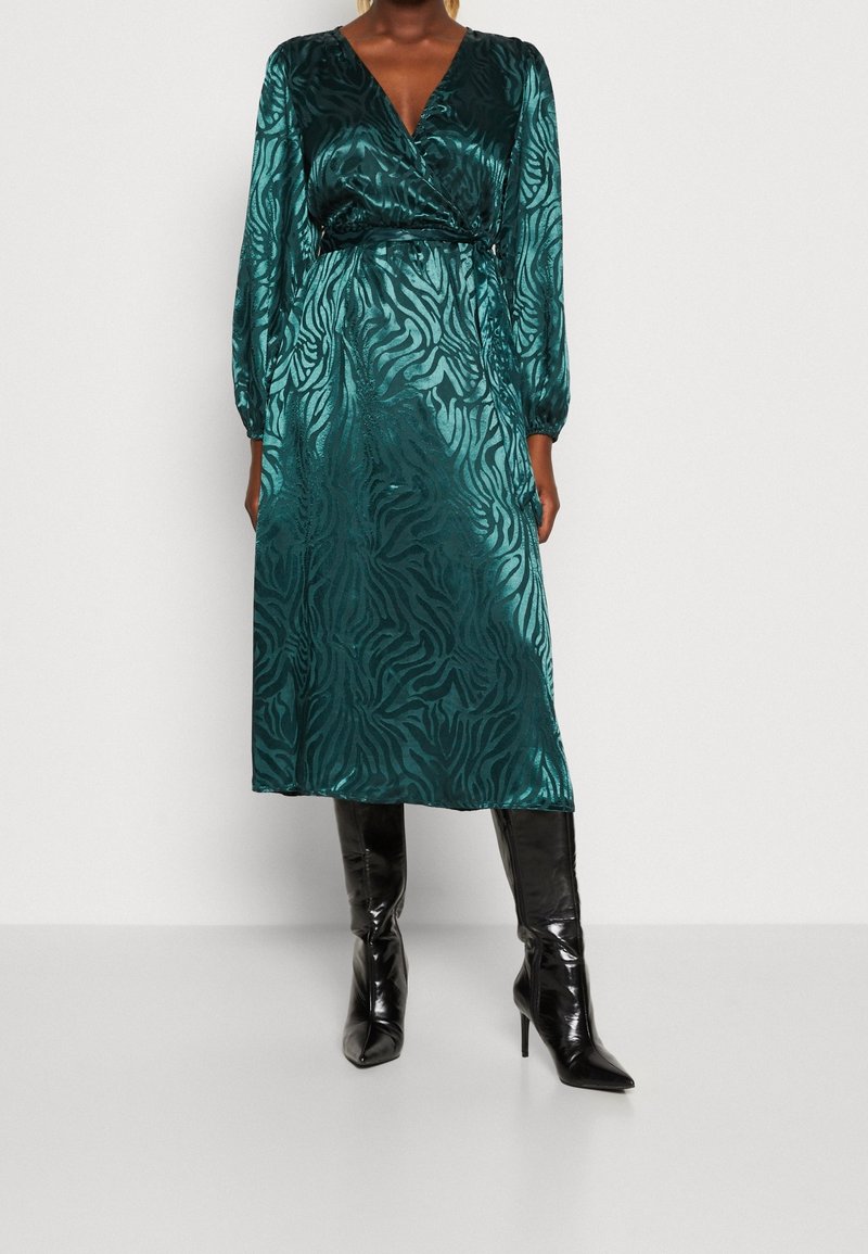 Femme portant une robe portefeuille en satin bleu sarcelle à imprimé zèbre et manches longues, associée à des bottes noires à talons hauts montant jusqu’aux genoux.