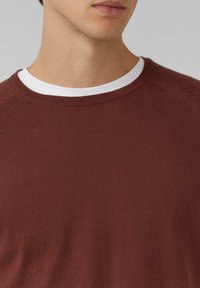 Zbli�żenie na osobę w brązowym swetrze robionym na drutach nałożonym na biały t-shirt z okrągłym dekoltem, pokazującą szyję i częściowo dolną część twarzy.