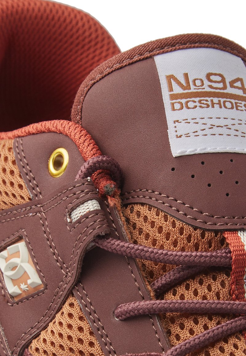 Chaussure de sport marron et orange dotée d'un mesh pour la respirabilité, cuir texturé, languette étiquetée, et œillets dorés.