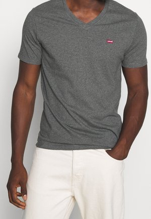 Homme portant un t-shirt ajusté gris foncé à col en V avec un petit logo rouge sur la poitrine et un pantalon clair, une main dans la poche.