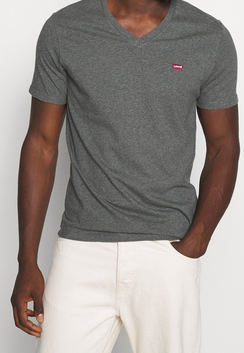 Homme portant un t-shirt ajusté gris foncé à col en V avec un petit logo rouge sur la poitrine et un pantalon clair, une main dans la poche.