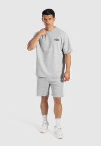 Camiseta de algodón gris y pantalones cortos a juego; corte relajado; logo en el pecho; usada con zapatillas blancas y calcetines deportivos. Iluminación uniforme, fondo neutro.