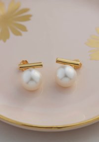 Orecchini a post in oro con grandi perle bianche lisce di forma rotonda. Esposti su un piatto rosa con accenti dorati.