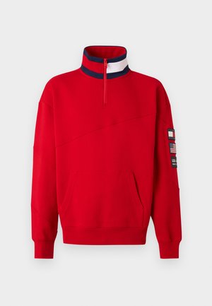 Rode quarter-zip trui met een kraag met marineblauwe en witte strepen, een voorvak en patches, waaronder een Amerikaanse vlag op de linker mouw.