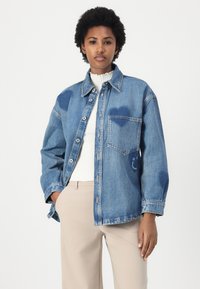 Denimjakke i blå, med knappestolpe, store lommer og hjerteformede detaljer i lysere blå. Kombinert med en ribbestrikket hvit topp og beige bukser.