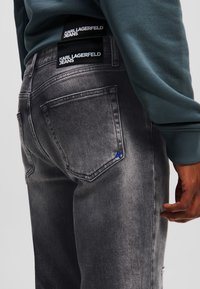 Jean gris de mezclilla con un acabado desgastado, con dos bolsillos traseros y un logo bordado en azul. Acompañado de un sudadera en azul verdoso.