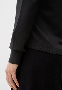 Linke Hand entspannt an der Seite, trägt einen langärmligen schwarzen Pullover mit gerippten Bündchen und einen passenden strukturierten schwarzen Rock vor einem schlichten Hintergrund.