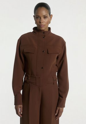 Vrouw met donker haar naar achter getrokken draagt een bruine jumpsuit met knopen en klepzakken en gouden oorringen, staand tegen een witte achtergrond.