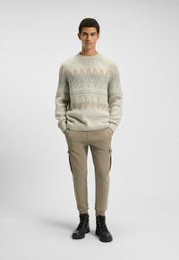 Cremefarbener Strickpullover mit blauen und braunen geometrischen Mustern, kombiniert mit beigen Cargo-Hosen und schwarzen Schnürstiefeln. Glatte Textur, lockere Passform.