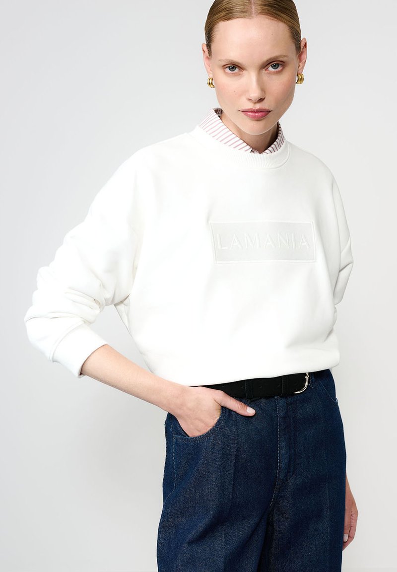 Biała bluza z logo "LAMANIA" wytłoczonym na przodzie, z ribbowanym okrągłym dekoltem, w zestawie z ciemnoniebieskimi spodniami z wysokim stanem. Gładka tekstura, luźny krój.