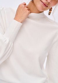 Biała sweter z wysokim kołnierzem i długimi, lekko bufiastymi rękawami. Miękka tekstura i prążkowane mankiety. Kolor jednolity, brak wzorów lub akcentów widocznych.