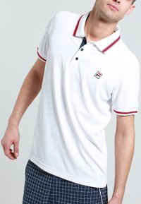 Vit polo-shirt med röda och marinblå randiga detaljer på kragen och ärmarna. Har en liten logotyp på bröstet. Slät textur, klassisk passform.