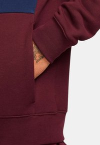 Felpa color bordeaux con un pannello blu sul petto, polsini a costine, tasca laterale e una texture liscia con dettagli di cucitura visibili.