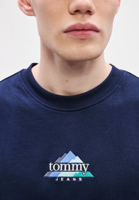 Tengerészkék pulóver bordázott nyakrésszel, kék és zöld árnyalatú hímzett hegylogóval, alatta "tommy JEANS" felirattal.
