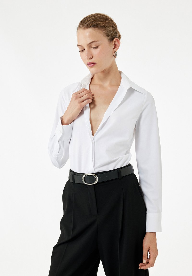 Vrouw die een wit knoopshirt en zwarte broek met een riem draagt, poseert met gesloten ogen en een hand bij de kraag.