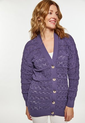 Frau lächelt, trägt eine violette gestrickte, geknöpfte Strickjacke mit Spitzenmuster und weiße Hose vor weißem Hintergrund.