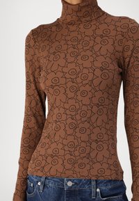 Brauner gerippter Rollkragenpullover mit schwarzem Blumenmuster, lange Ärmel und taillierte Passform, getragen mit blauen Jeans.