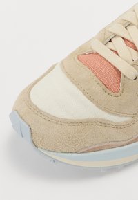 Beige sneakers med overdel i semsket skinn og mesh, kontrastfarget korallpanel, teksturert såle og lyseblå detaljer.