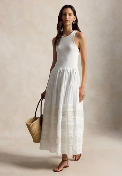 Naine, kes kannab varrukateta valget kleiti tekstuurse seelikuga, hoiab käes punutud roost kotti ja kannab pruune lameda tallaga sandaale.