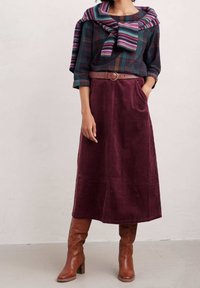 Camicia a quadri con tonalità verdi, borgogna e blu, abbinata a una gonna in corduroy borgogna e stivali alti fino al ginocchio marroni con tacco. Sciarpa di lana annodata intorno alle spalle.