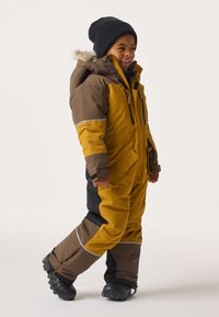 Jong kind dat glimlacht, wandelend in een bruine en mosterdgele wintersneeuwpak met zwarte laarzen en een zwarte beanie op een witte achtergrond.