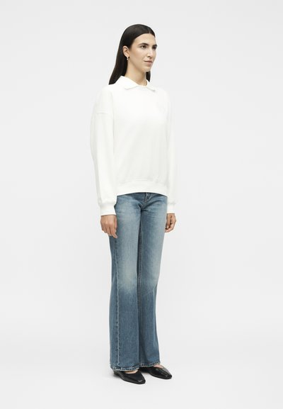Femme debout, portant une chemise blanche à manches longues avec col, un jean évasé bleu clair et des chaussures noires pointues sur un fond blanc uni.