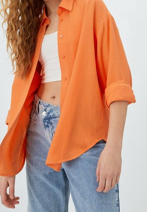 Persoon die een open oranje overhemd draagt over een witte crop top en lichtblauwe high-waisted jeans tegen een effen achtergrond.
