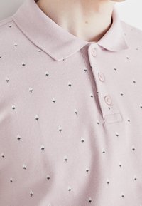 Pánská polo košile v světle růžové barvě z bavlněného úpletu, zdobená malými černobílými geometrickými vzory, s knoflíkovou légou na tři knoflíky a krátkým límcem.