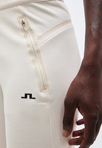 Nahaufnahme einer Hand neben weißen Hosen mit einem senkrechten beigen Reißverschluss und einem kleinen schwarzen geometrischen Logo.